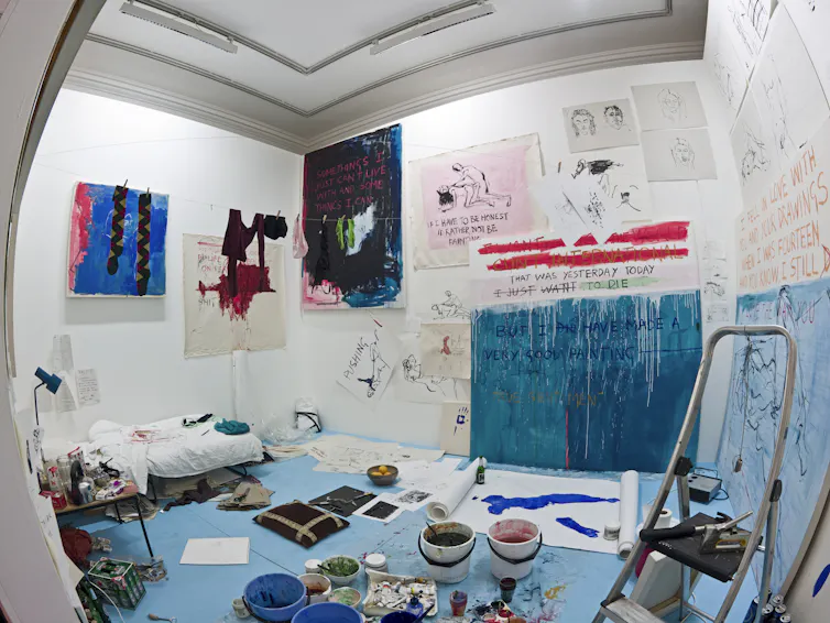 An artist's room with a bed and plentiful art supplies and paintings on the walls