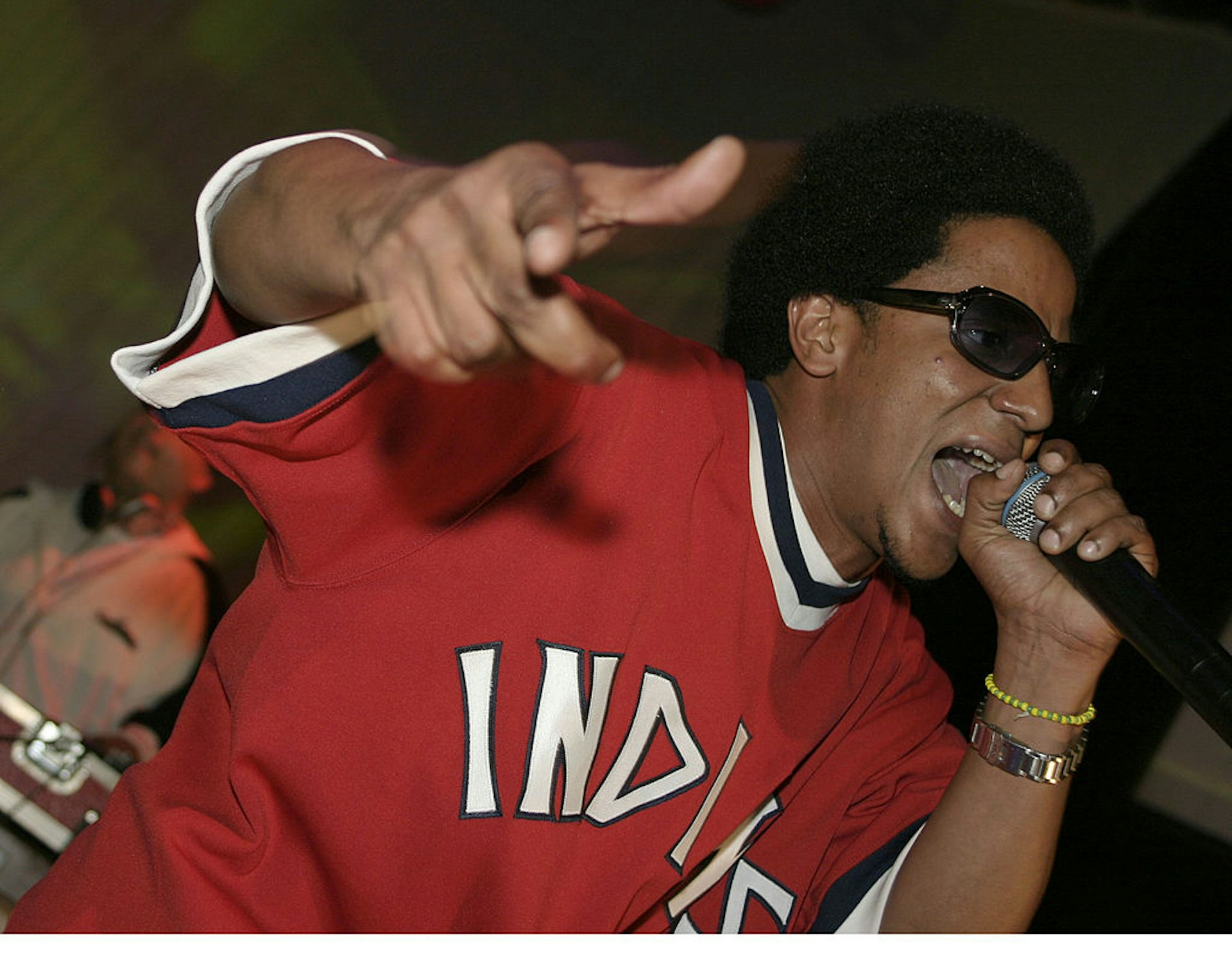 Man with afro wearing sunglasses and a red baseball jersey gestures while rapping into a microphone.