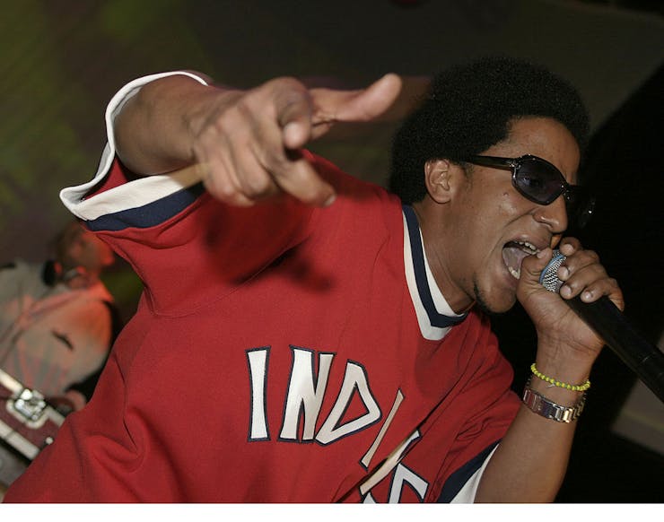 Man with afro wearing sunglasses and a red baseball jersey gestures while rapping into a microphone.