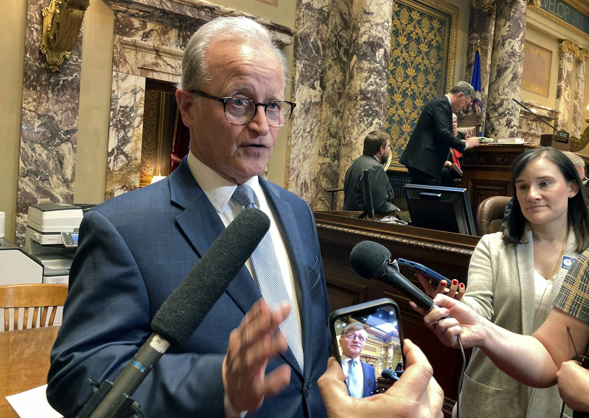 Standing in an ornate chamber, a man in a tie talks to reporters who hold microphones and cellphones up to his face.