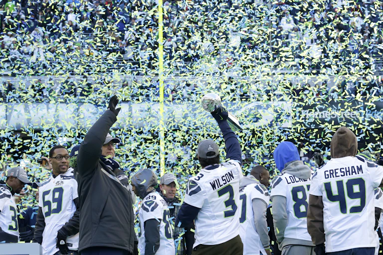 Man in a gray jacket waves, while surrounded by confetti and football players.