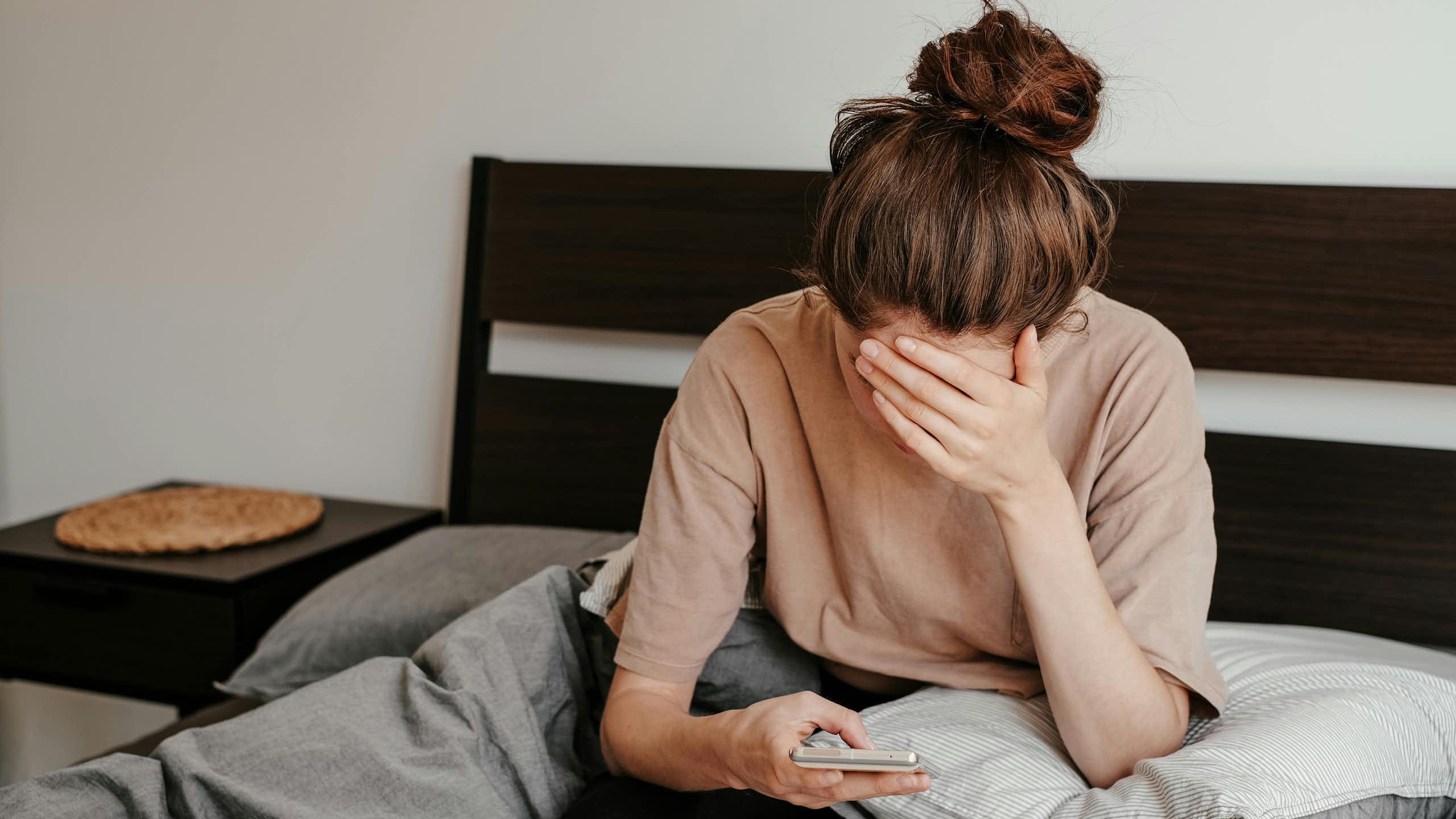 A woman sitting in bed resting her forehead against her hand as she scrolls on her phone