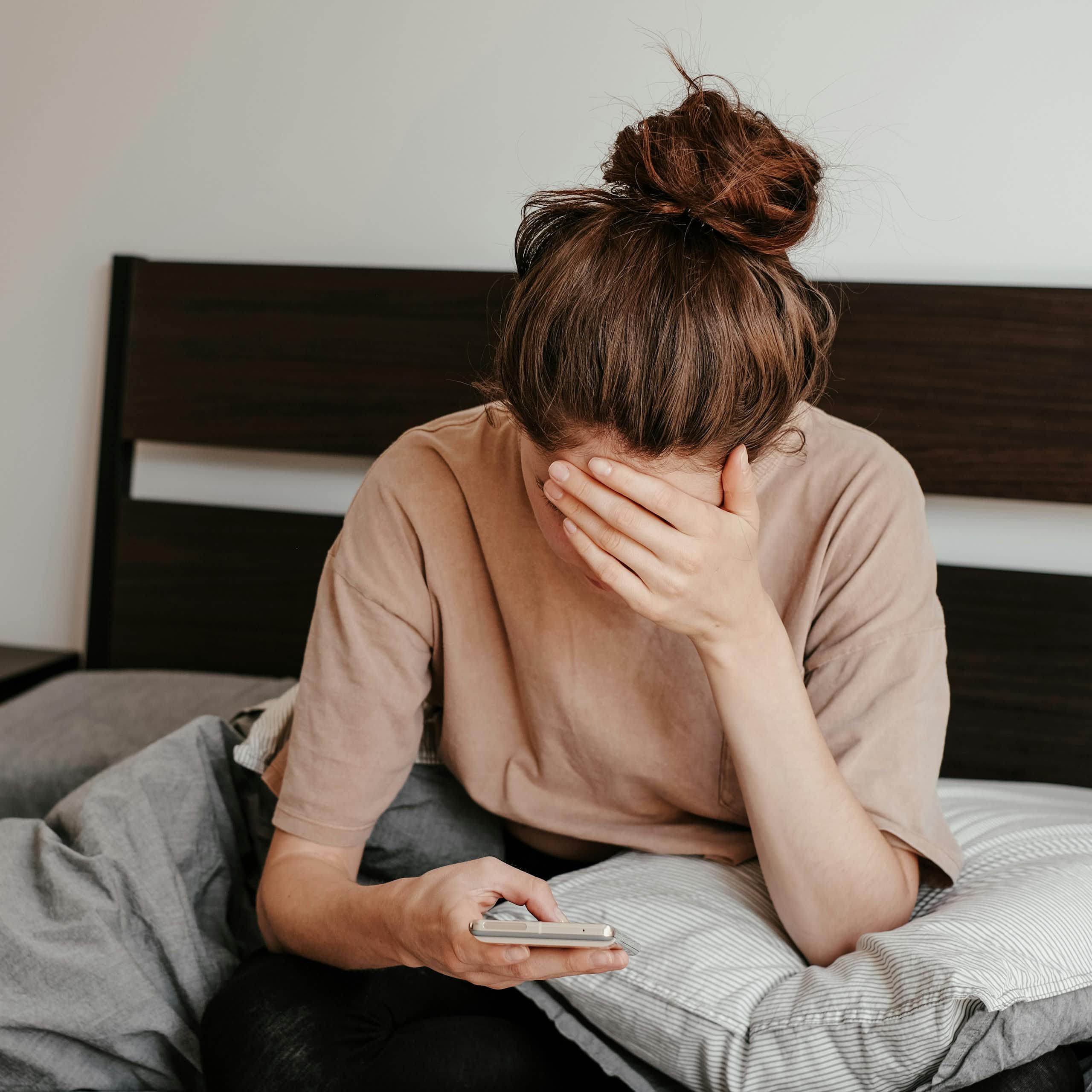 A woman sitting in bed resting her forehead against her hand as she scrolls on her phone