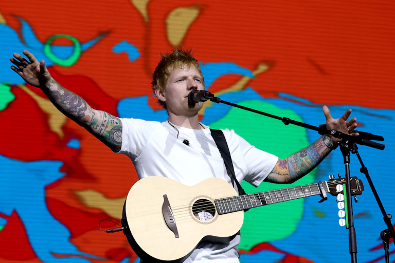 Pop star Ed Sheeran in a white t-shirt, holding a guitar and with a colourful background.