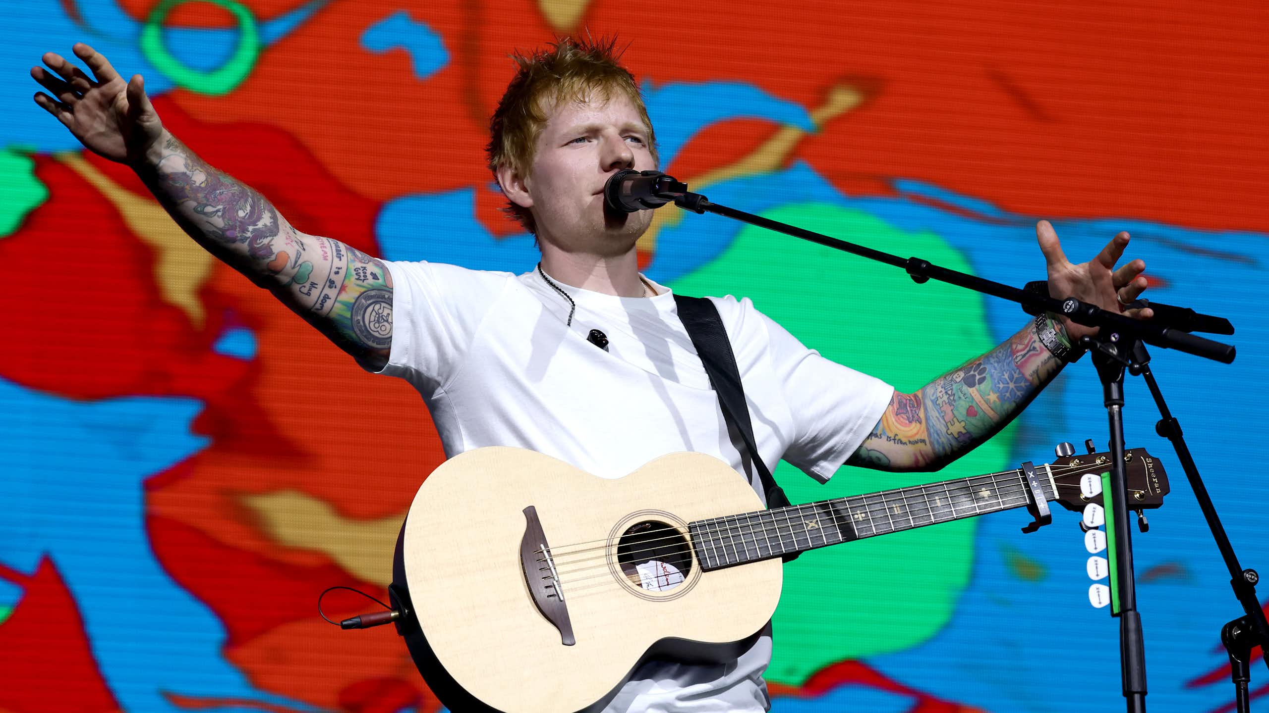 Pop star Ed Sheeran in a white t-shirt, holding a guitar and with a colourful background.