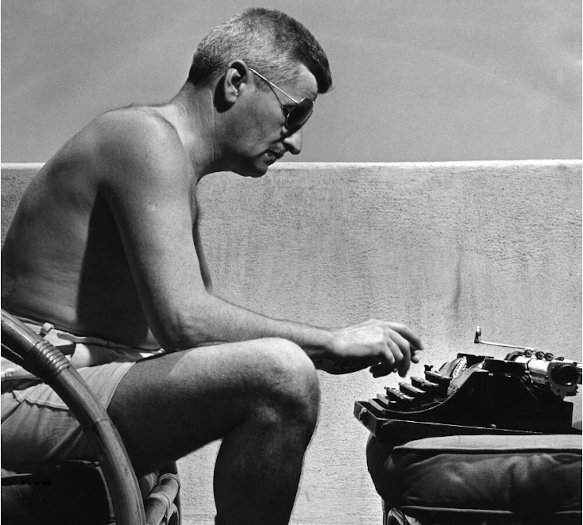 A shirtless man wearing shorts and sunglasses sitting at this typewriter in the sun.