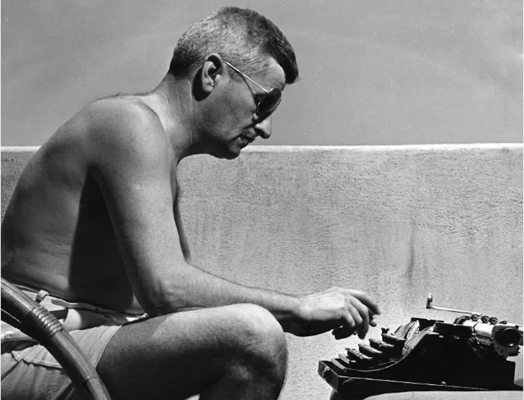 A shirtless man wearing shorts and sunglasses sitting at this typewriter in the sun.
