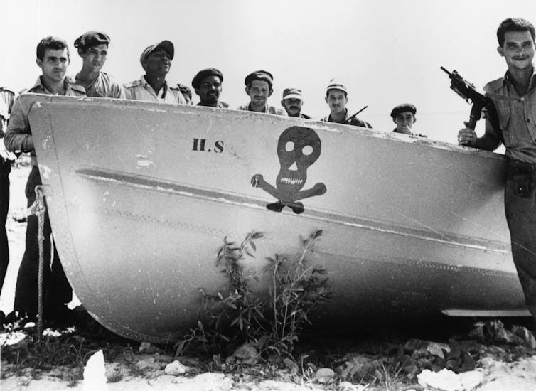 A group of men with guns stand by a boat with a skull and crossbones motif.