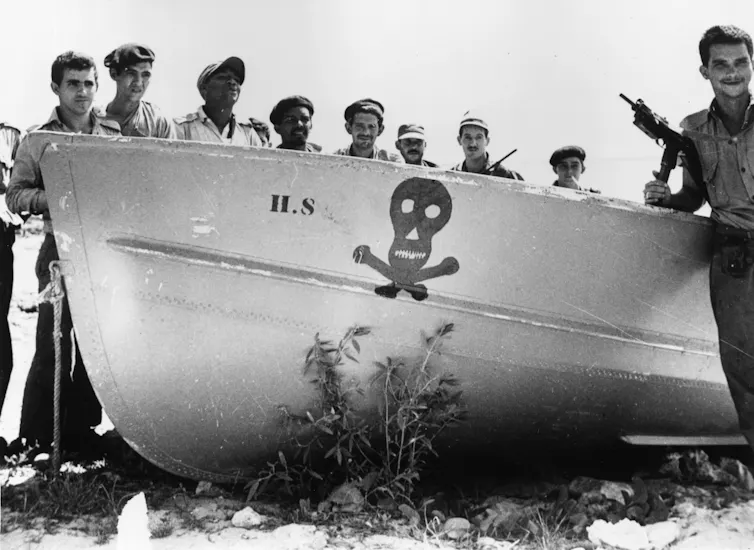 A group of men with guns stand by a boat with a skull and crossbones motif.