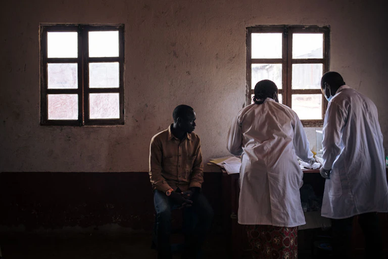 Patient sitting in chair while two people in white coats hunch over a table of medical and research supplies