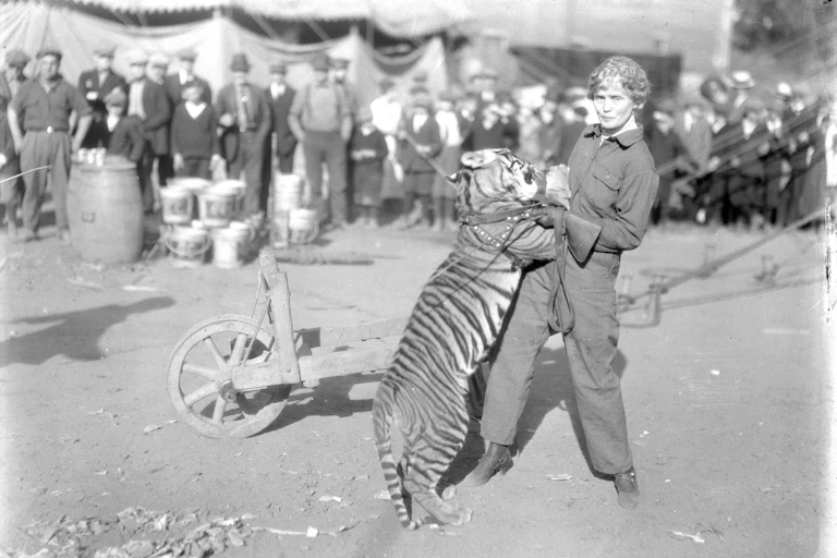 Black and white photo of a young woman striking a defiant pose while supporting a tiger standing on its hind legs.