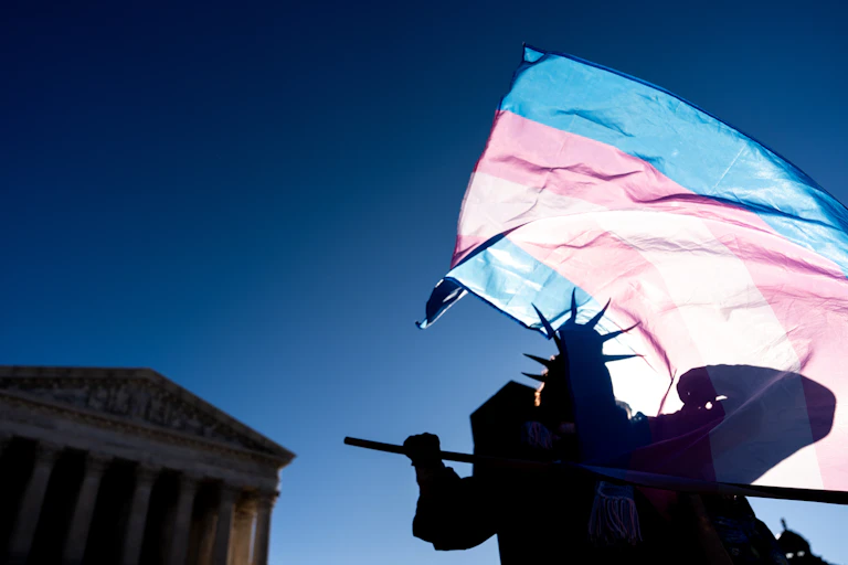 Silhouette of person dressed as Statue of Liberty holding a transgender flag up against a blue sky, the Supreme Court looming in distance