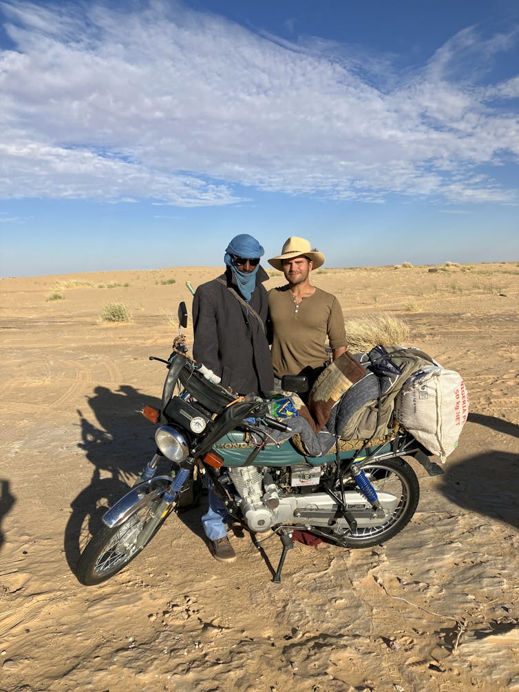 A man wearing a dark jacket and a blue headscarf with a motorbike, standing next to a man in a cowboy hat.