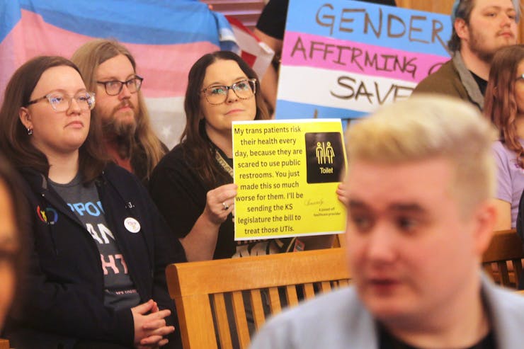 People holding signs in protest, one reading 'My trans patients risk their health every day, because they are scared to use public restrooms. You just made this so much worse for them. I'll be sending the KS legislature the bill to treat those UTIs'