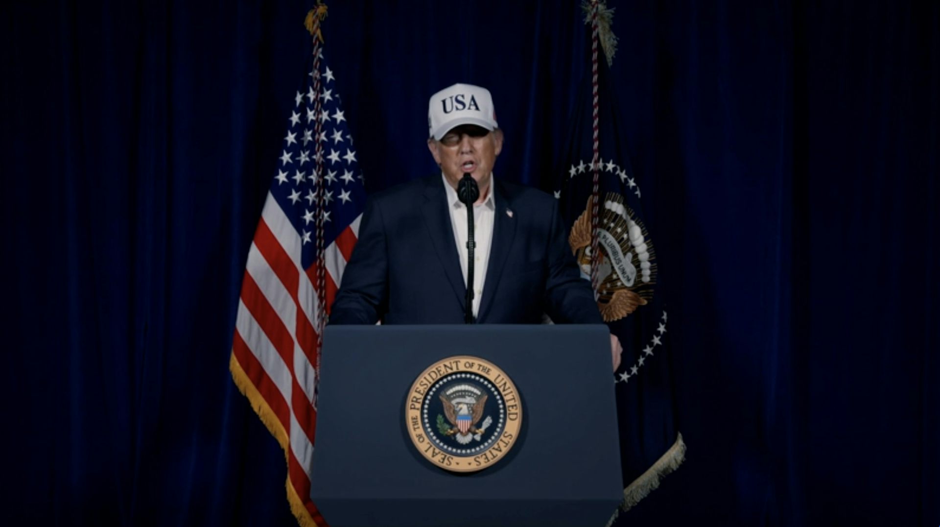 A man in a suit and a baseball cap with USA on it stands at a podium.