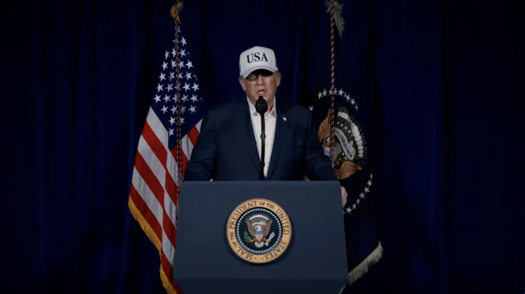 A man in a suit and a baseball cap with USA on it stands at a podium.