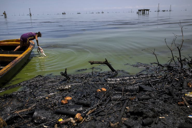 A person washes a fish out of murky water near shore where an oil slick covers debris.