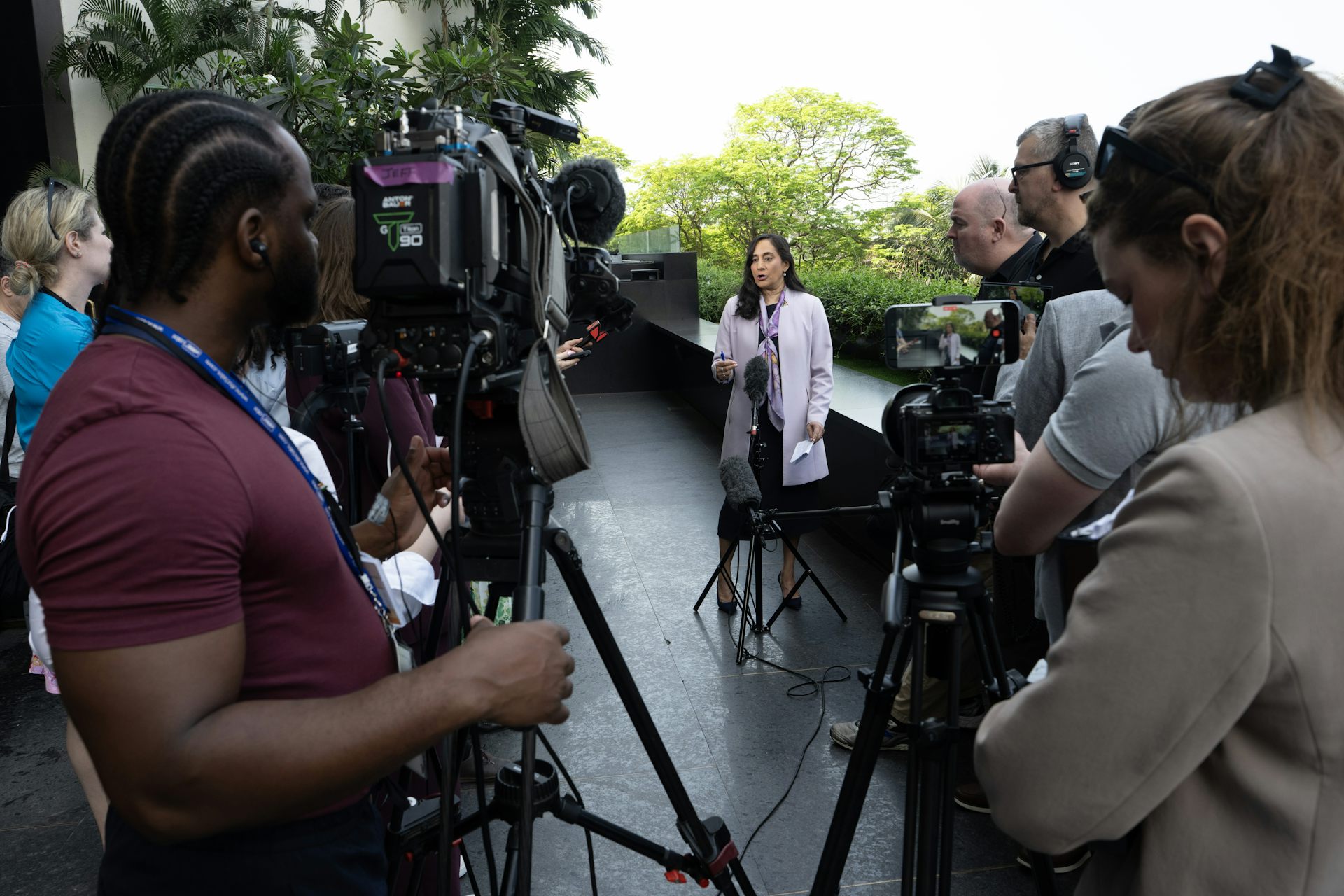 Una mujer se pone de pie y habla con un grupo de reporteros y camarógrafos en un área abierta.