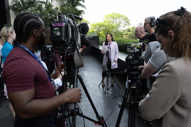 Una mujer se pone de pie y habla con un grupo de reporteros y camarógrafos en un área abierta.