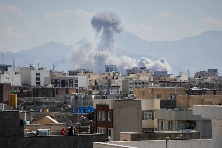 A large plume of smoke over a city as people watch from rooftops.
