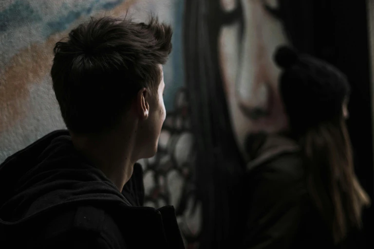A male youth looks on as a young woman spray paints a dark building.