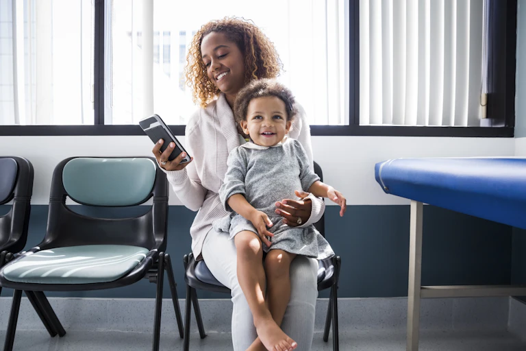Woman in waiting room holding a toddler looks at her smartphone.