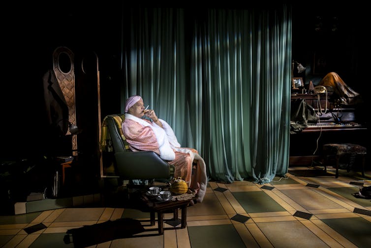 Production image: a man sits in a pink dressing gown, smoking.