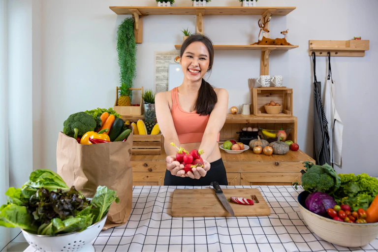 Happy Asian woman holds veg standing in kitchen with shelves, colourful fruit and veg on display