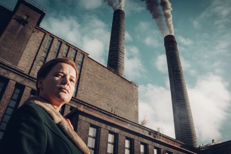 A still from the Netflix series, Lead Children, in which a woman stands outside of an industrial factory which is bellowing out plumes of smoke.