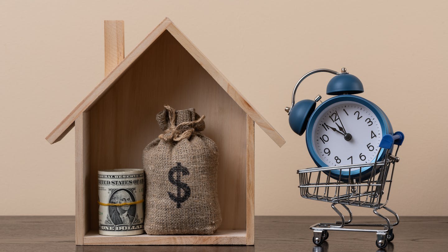 A symbolic wooden house, containg a stack of $1 bills and a money bag with a dollar symbol, sits next to an alarm clock in a grocery cart.