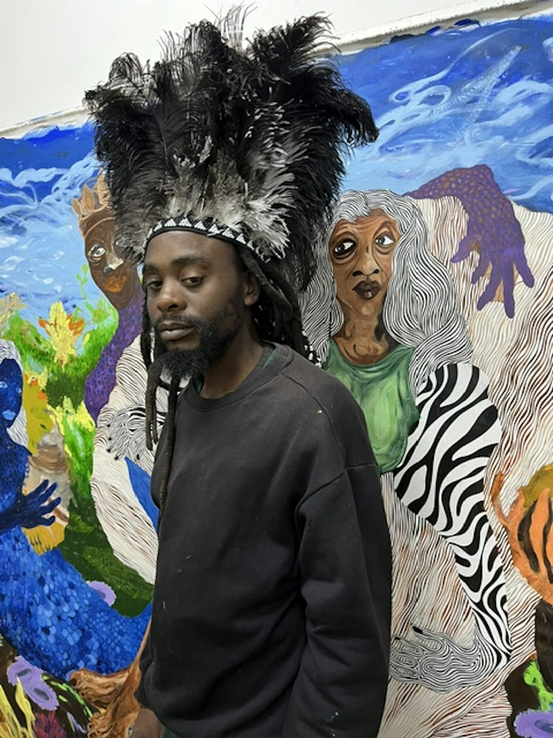 A young African man with dreadlocks stands in front of a colourful painting wearing an elaborate feathered headpiece.