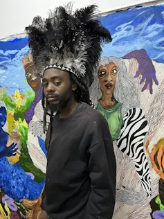 A young African man with dreadlocks stands in front of a colourful painting wearing an elaborate feathered headpiece.