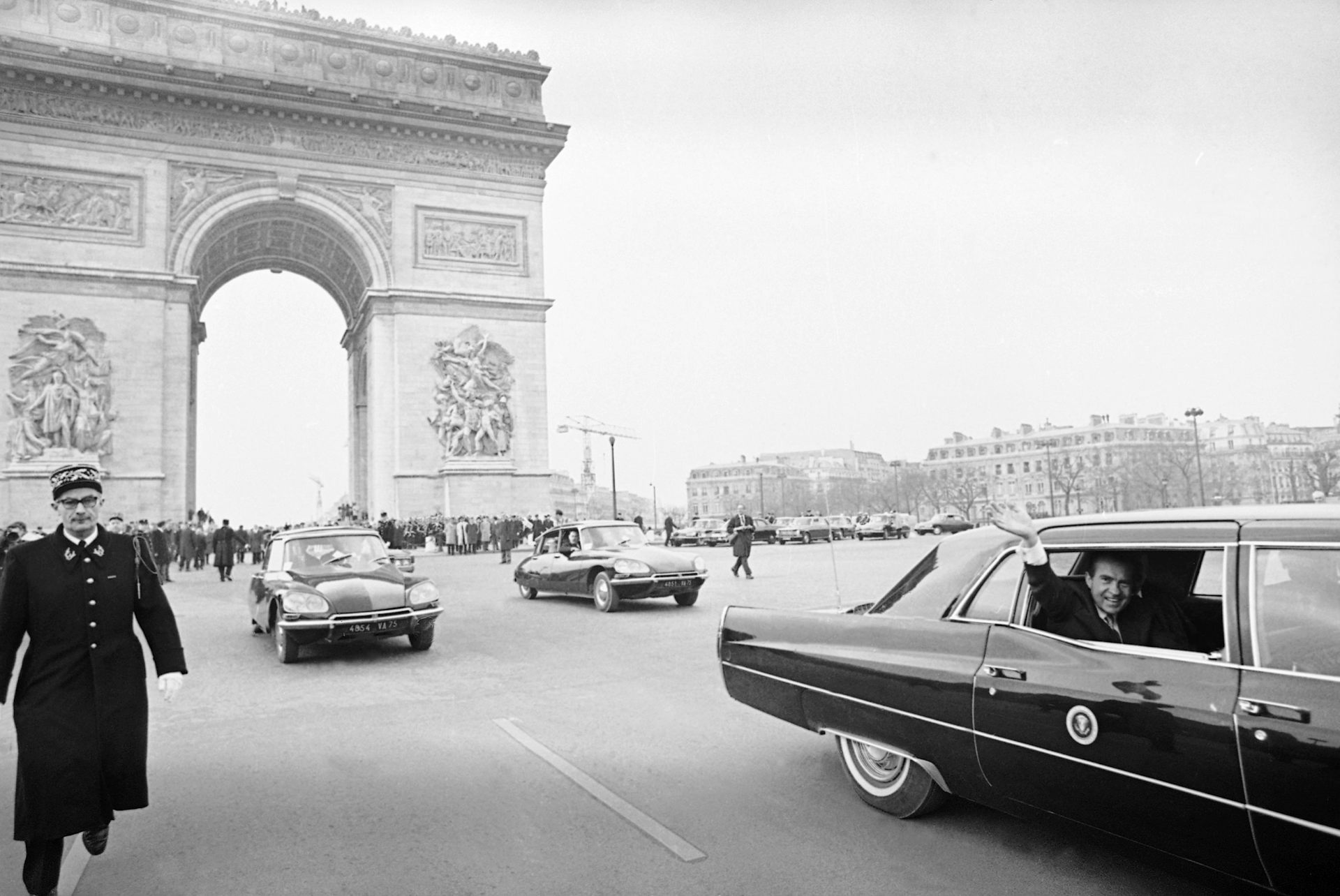 A man in a 1960s style car waves from a back window near a triumphal arch and a uniformed police officer in a black and white photo.
