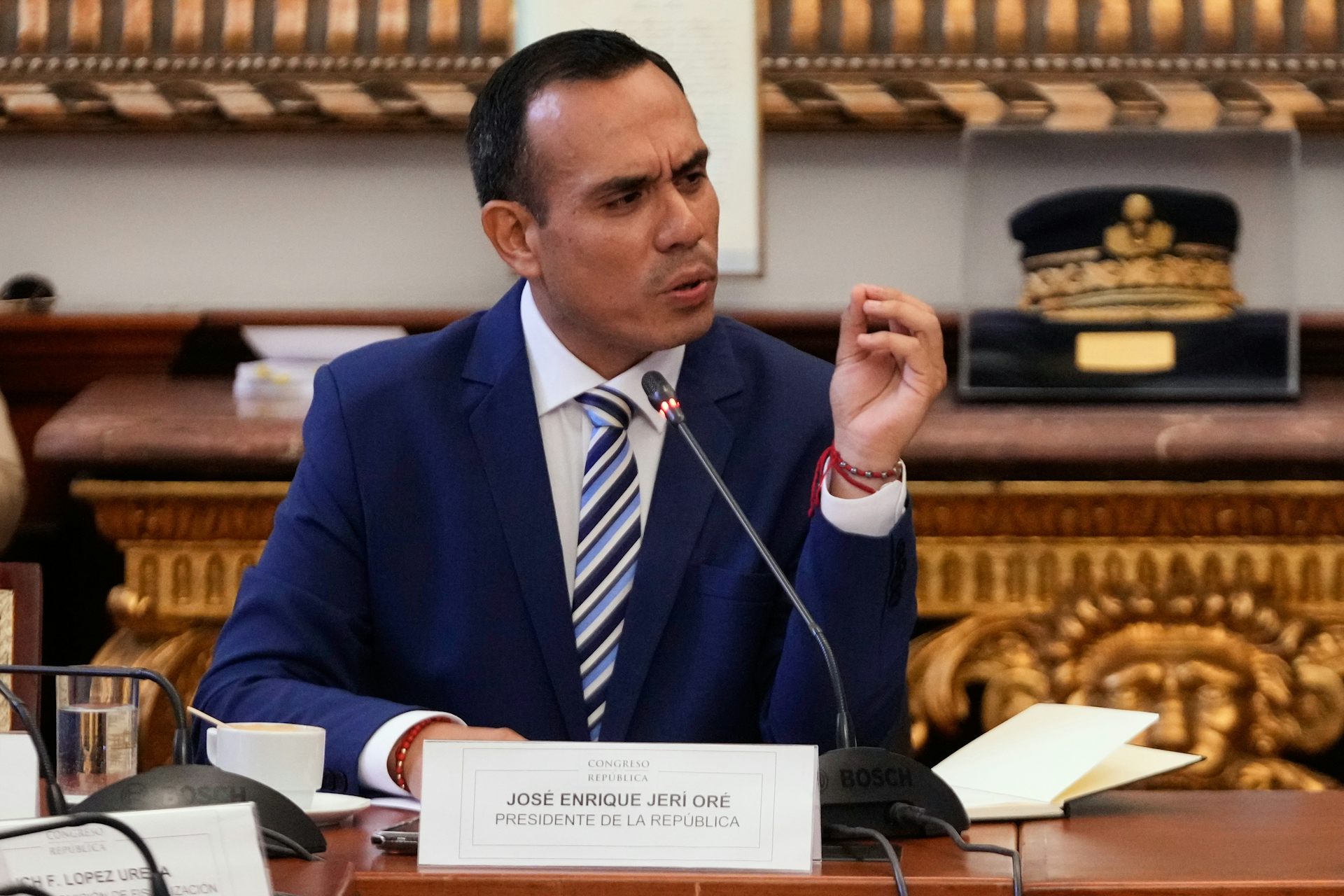 Un hombre de mediana edad con un traje azul oscuro se sienta en un escritorio y habla por un micrófono.