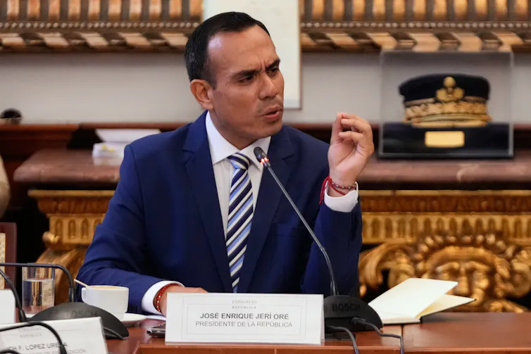 Un hombre de mediana edad con un traje azul oscuro se sienta en un escritorio y habla por un micrófono.