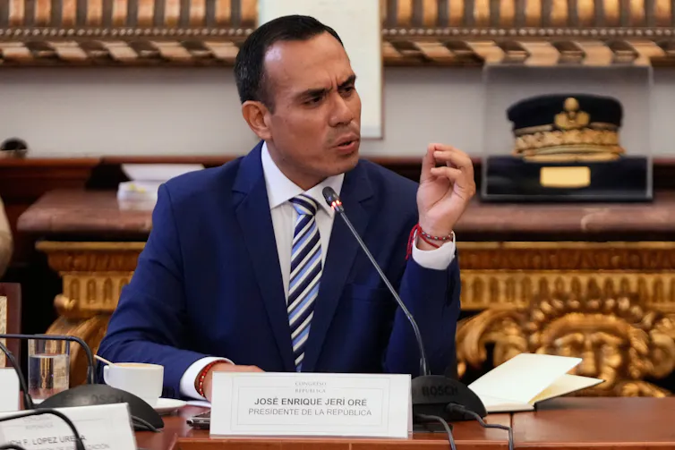 A middle-aged man in a dark blue suit sits at a table speaking into a microphone