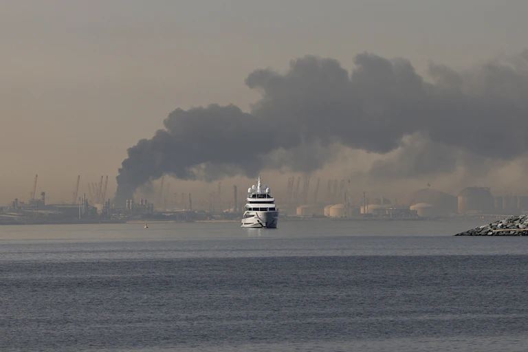 a white yacht sails forward in the open water as dark smoke billows in the skyline in the background