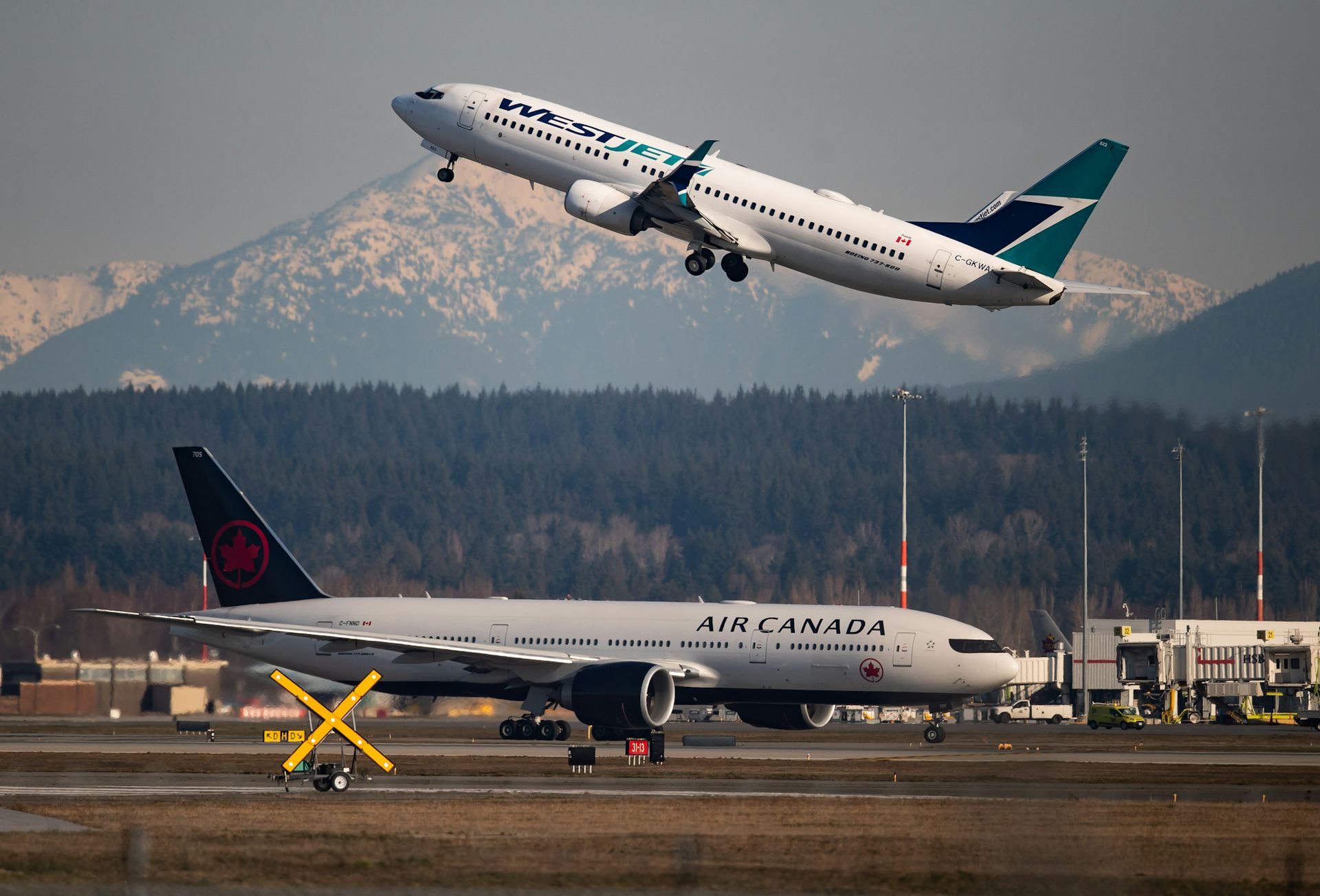 An airport tarmac with a plane taxiing in the foreground and one taking off in the background