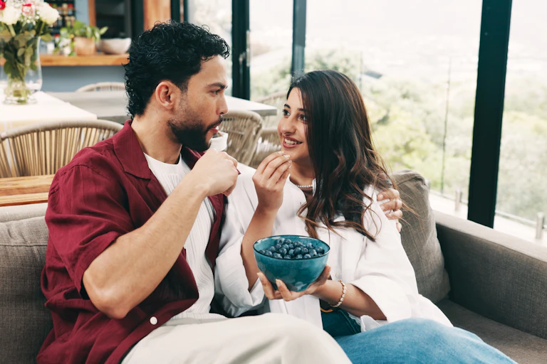 A couple sitting next to each other on a sofa, sharing a bowl of blueberries.