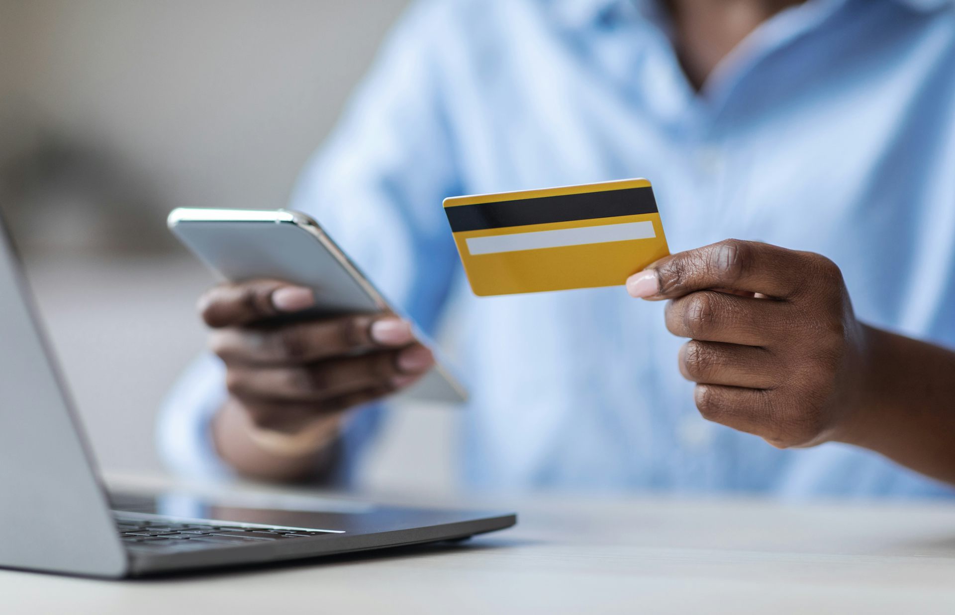 Woman's hands holding a bank card and a mobile phone; she is seated at a laptop