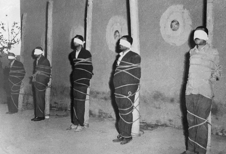 Five men are blindfolded and bound against posts.