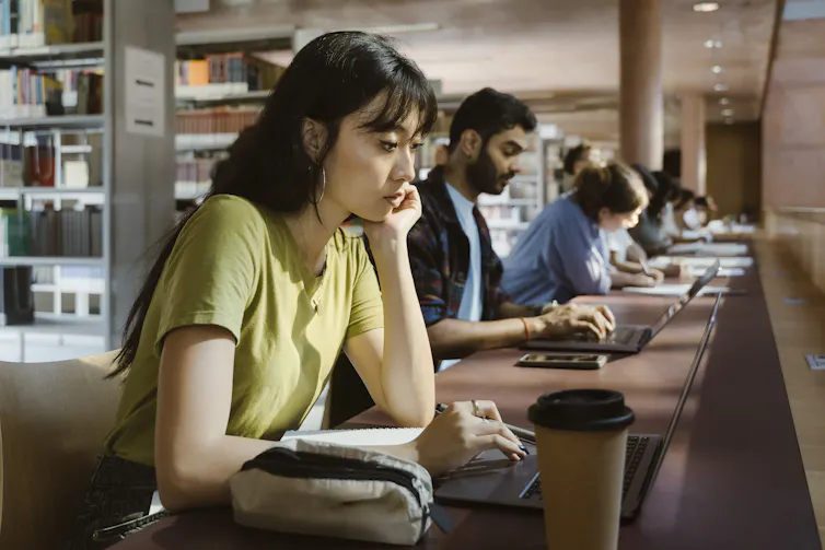 An image shows a row of college-aged people sitting at a long desk in a library.