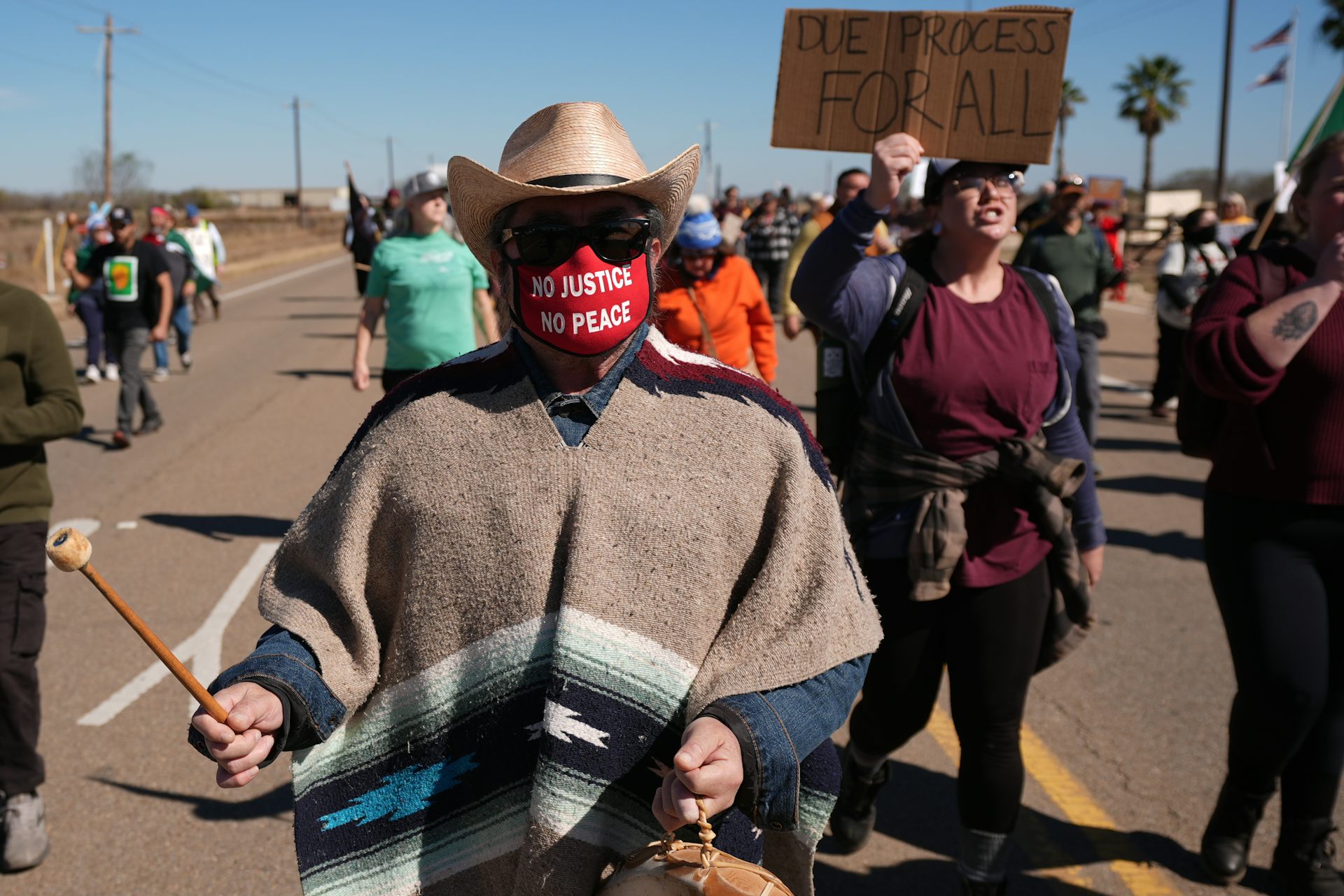 A man wearing a poncho and a mask that says 'no justice, no peace' bangs on a drum during a protest.