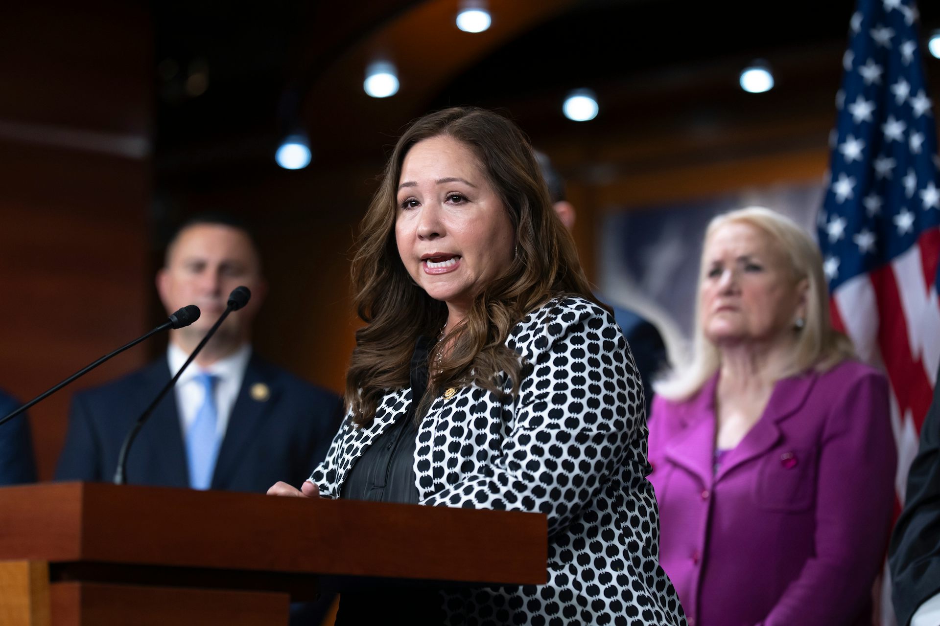 A female member of Congress in a black-and-white polka dotted jackets stands at a lectern and speaks during a news conference.