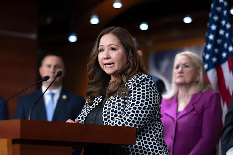 A female member of Congress in a black-and-white polka dotted jackets stands at a lectern and speaks during a news conference.