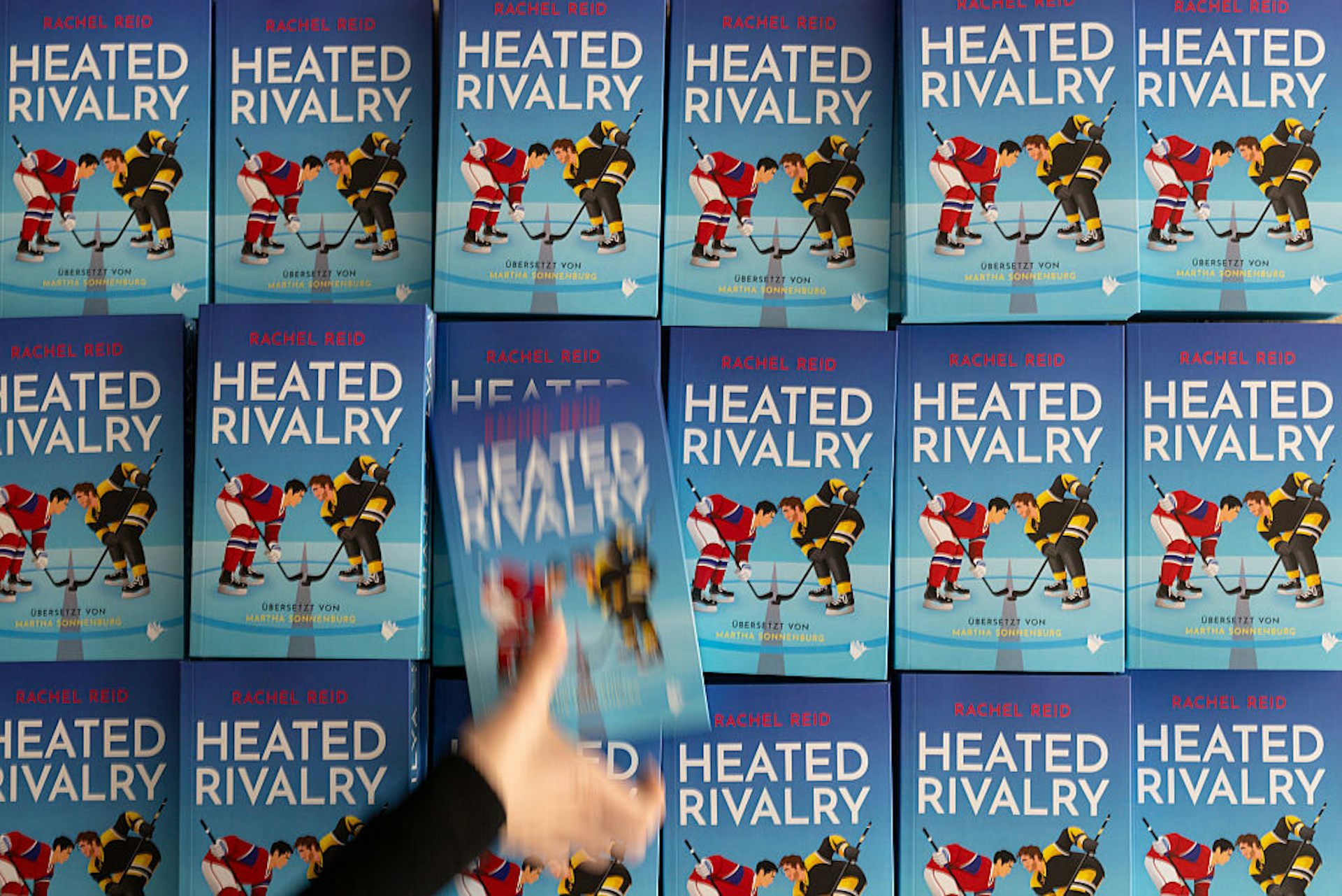 A blurry hand-in-motion removes a book from a display featuring the same title, 'Heated Rivalry.'