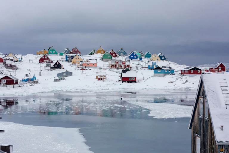 A snowy landscape dotted with colourful houses with an icy shoreline in the foreground.