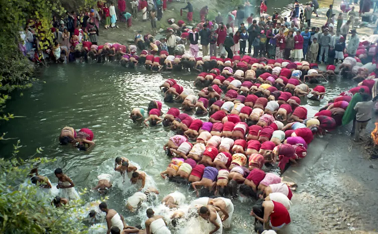 Several women draped in red or white cloth bathe in the river, while men dressed in white also take part in the ritual dip.