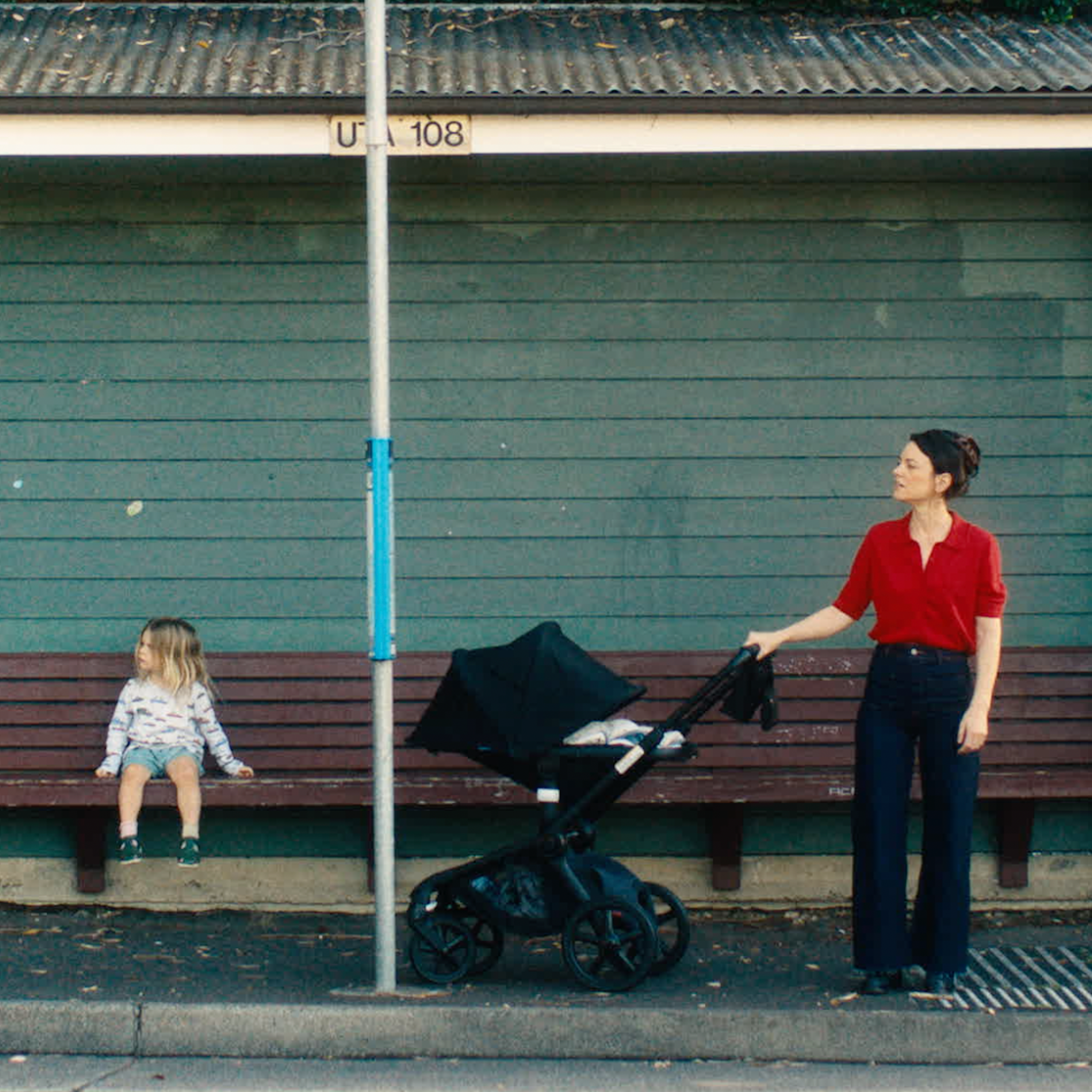 Film still: a mother and a toddler at a bus stop.