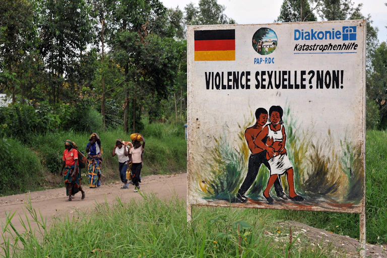 Women walk on a dirt road past a sign with an illustration of a man accosting a woman.
